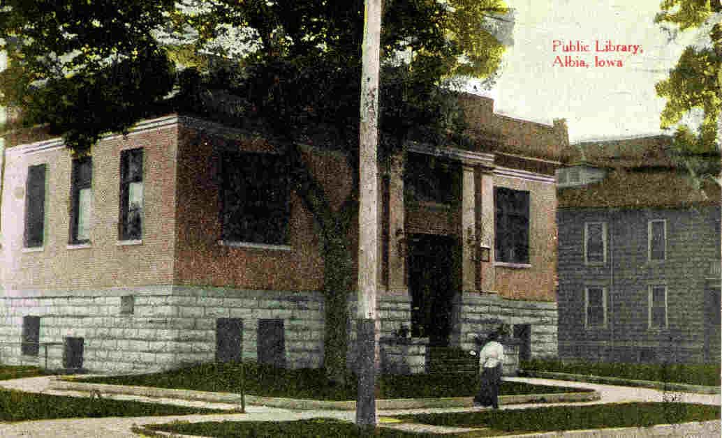 Albia postcard 1. Color postcard showing the of the Albia Public Library and person walking on sidewalk. Streetlight and tree partially blocking view of the building. Text reads "Public Library, Albia, Iowa" Source: unknown.