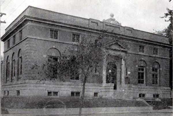 Black and white image showing the front exterior of the Oskaloosa Public Library. Tree partially blocking view of the building. 