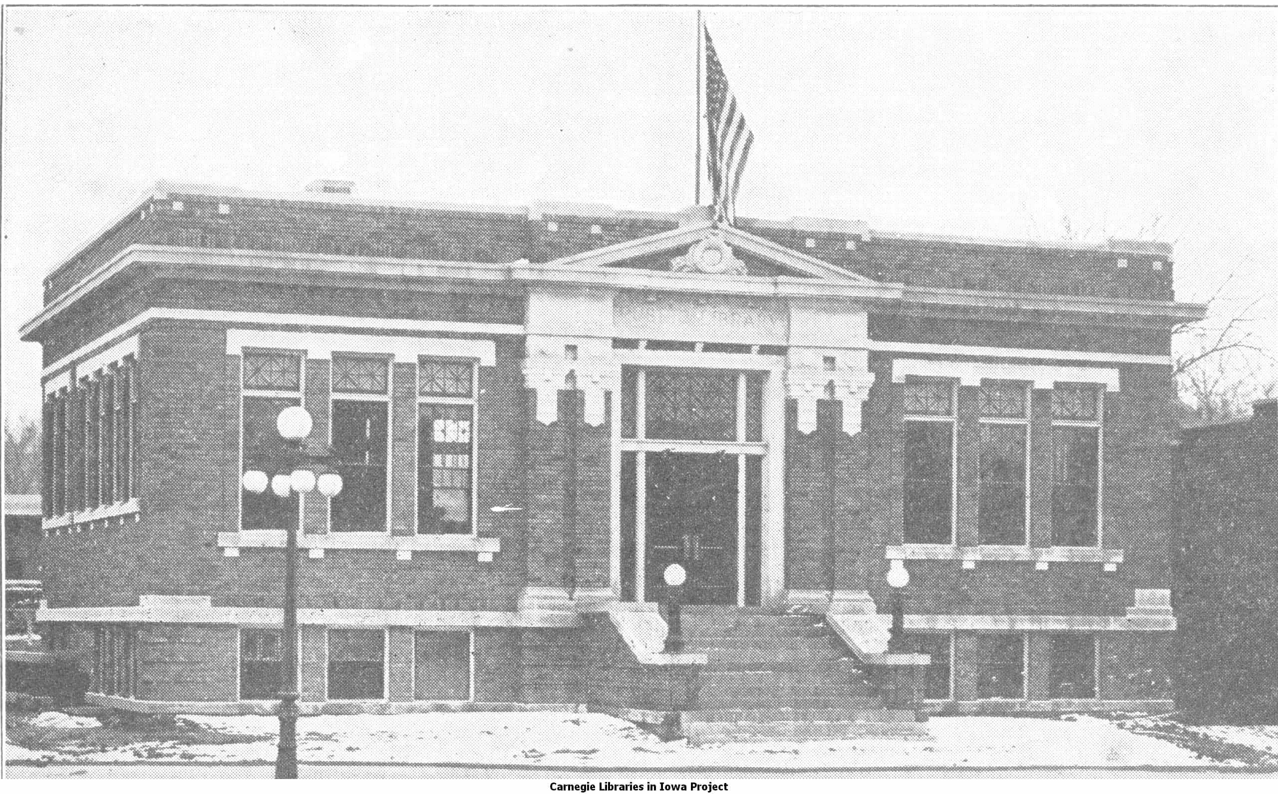 Black and white image showing the front exterior of the Traer Public Library. An American flag is also visible. Taken from the 8th Report of the Iowa Library Commission, coverage 1914-1916. (Published in 1916), page 12.