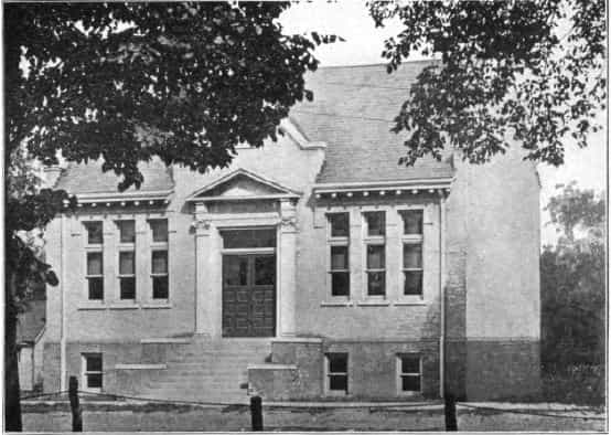 Black and white image depicting the front exterior of the Lake City Public Library. Trees are partially blocking the roof of the library. Taken from the 5th Report of the Iowa Library Commission, coverage 1908-1910. (Published in 1909), unnumbered page between pages 16-17.