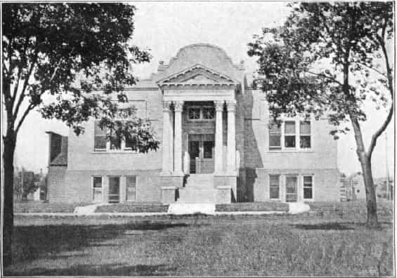 Black and white image showing the front exterior of the Rockwell City Public Library. Trees are partially blocking parts of the image. Taken from the 5th Report of the Iowa Library Commission, coverage 1908-1910. (Published in 1909), unnumbered page between pages 16-17.