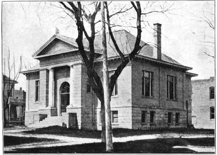 Black and white image showing the front and side exterior of the Sheldon Public Library. Tree and lamppost blocking center of image. 
