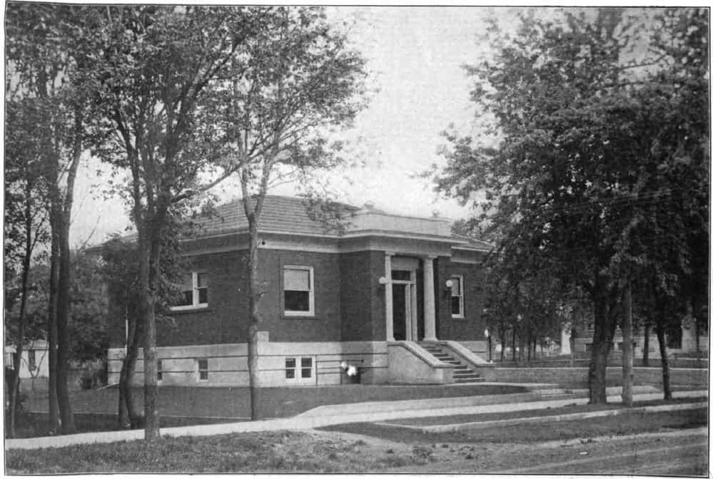 Black and white image showing the front and side exterior of the Sanborn Public Library. Trees partially blocking view of the building. Taken from the 6th Report of the Iowa Library Commission, coverage 1910-1912. (Published in 1913), opposite page 16.