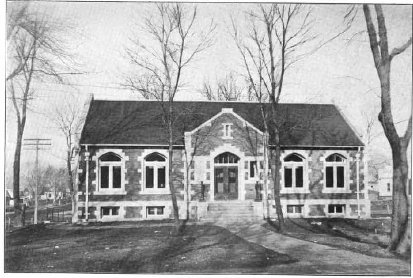 Black and white image showing the front exterior of the Audubon Public Library. Trees partially blocking view of the building. Taken from the 6th Report of the Iowa Library Commission, coverage 1912-1914. (Published in 1914), unnumbered page opposite [page 7]. 