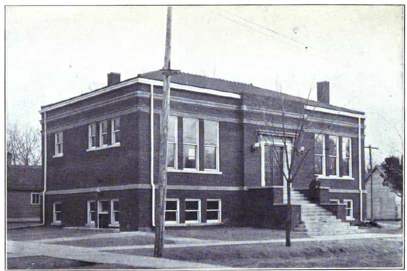 Black and white image showing the front and side exterior of the Dunlap Public Library. Lamppost and tree partially blocking view of the building. Taken from the 7th Report of the Iowa Library Commission, coverage 1912-1914. (Published in 1914), opposite page 19.