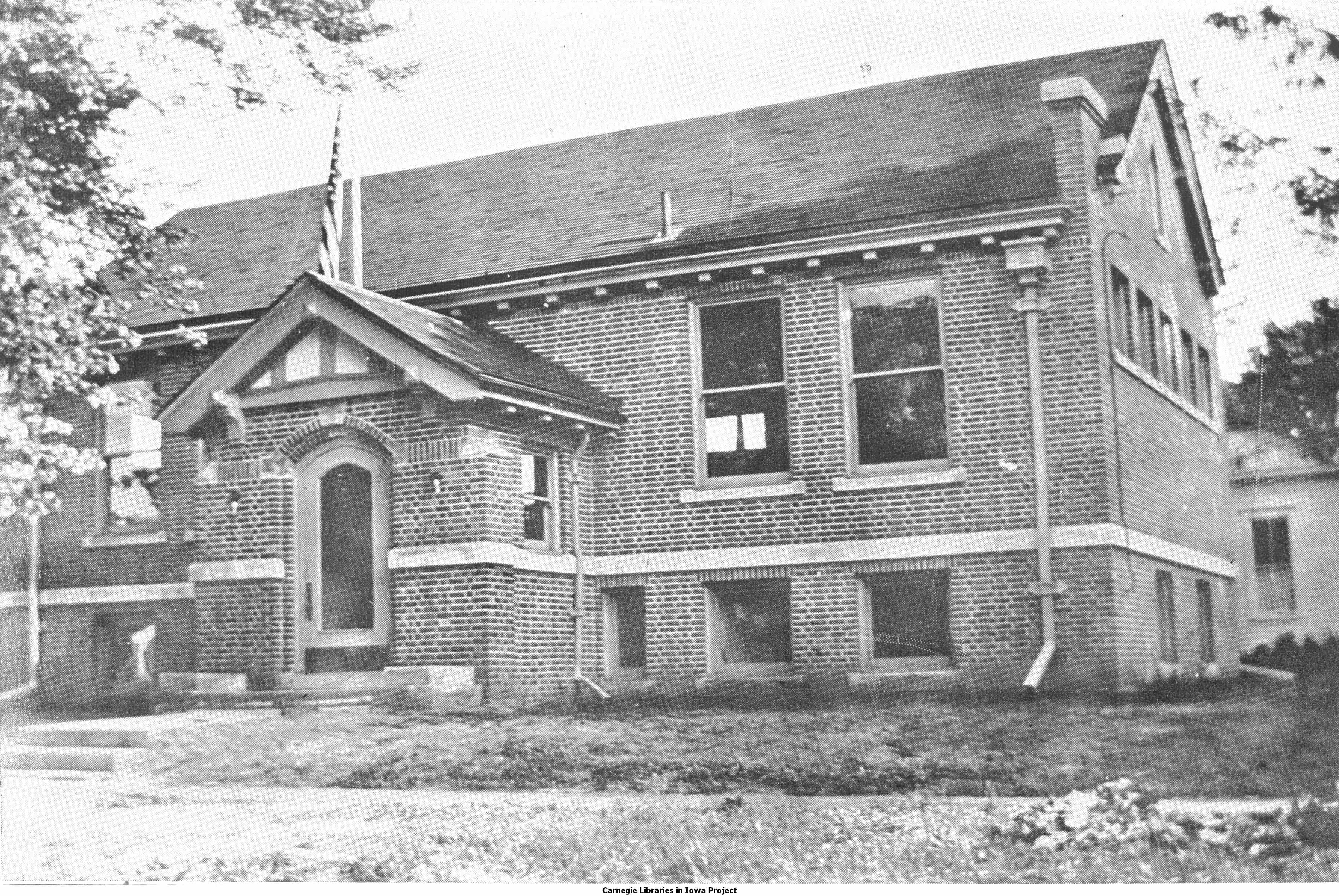 Black and white image showing the front exterior of the Colfax Public Library. Trees partially blocking view of the building. 