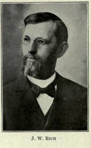 A bust photograph of Joseph Rich. He is wearing a suit and bowtie. He has a beard, hair slicked back with a side part, and small round glasses.