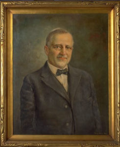 A framed painting of Bohumil Shimek. He is wearing a suit and bowtie. He has short white hair, a short white beard, and is wearing round glasses.