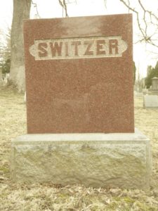 A full color photograph of Margaret Switzer's tombstone. It is red granite with the name Switzer inscribed into it.