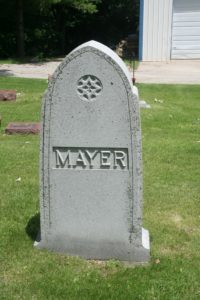 A close up color photograph of the Mayer family head stone. It is a pointed oval shape made of gray stone. The Mayer name is carved into it along with a decorative circular carving.