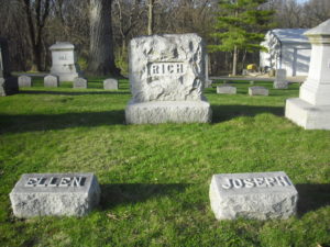 A color photograph of the grave of Joseph Rich. An upright gray stone headstone in the background says rich. There are two flat gray stone markers in front. The one on the left says Ellen and the one on the right says Joseph.