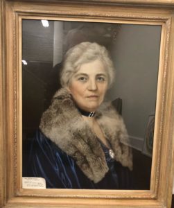 A framed portrait of Mrs. F. L. Maytag wearing a blue dress and blue coat with a fur shawl. She has a thick black necklace with a silver pendant.